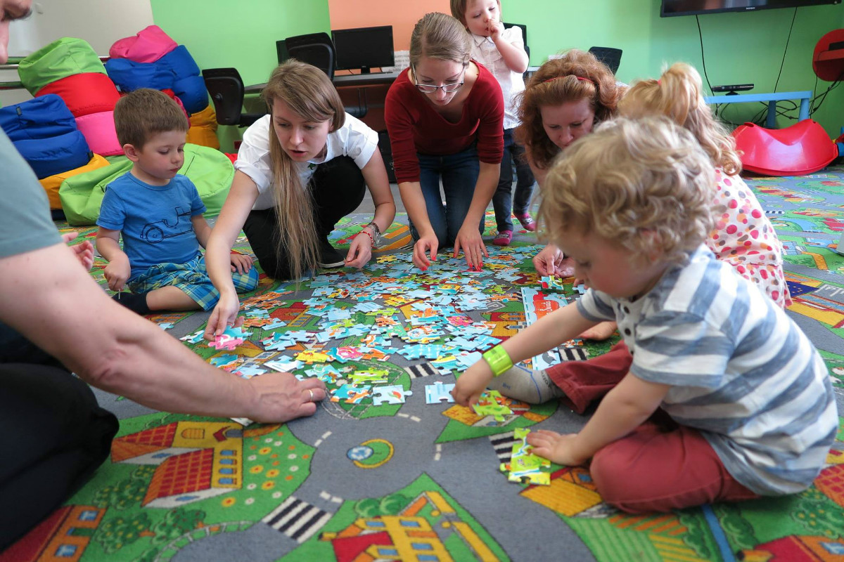 Wnętrze biblioteki.  Na kolorowej wykładzinie dzieci z opiekunami układają puzzle.
