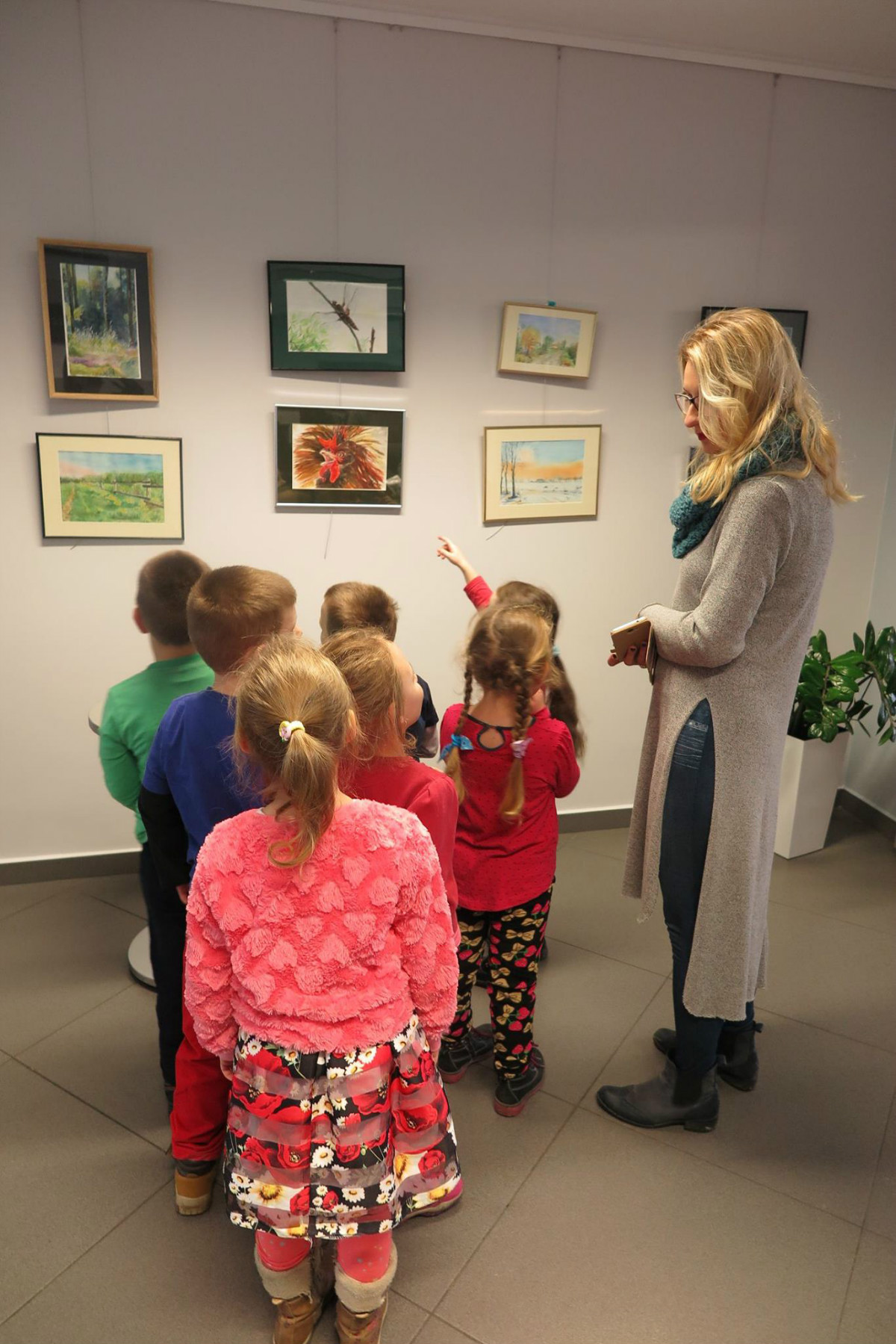 Wnętrze biblioteki,mediateka. Grupa dzieci z opiekunką ogląda wystawę akwareli Hanny Iwanów.