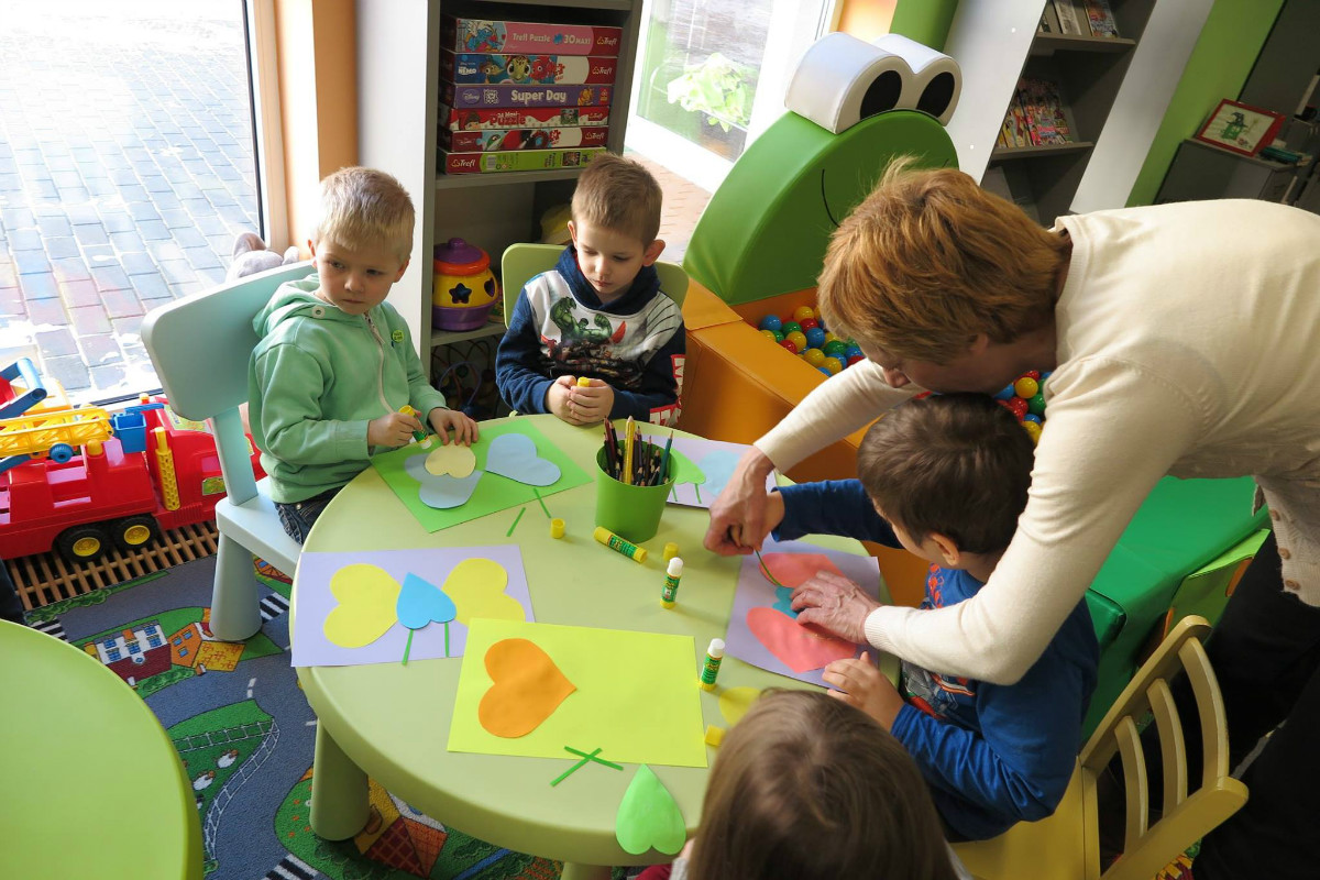 Przy stoliku siedzą dzieci z opiekunem razem wykonują prace plastyczne. 
