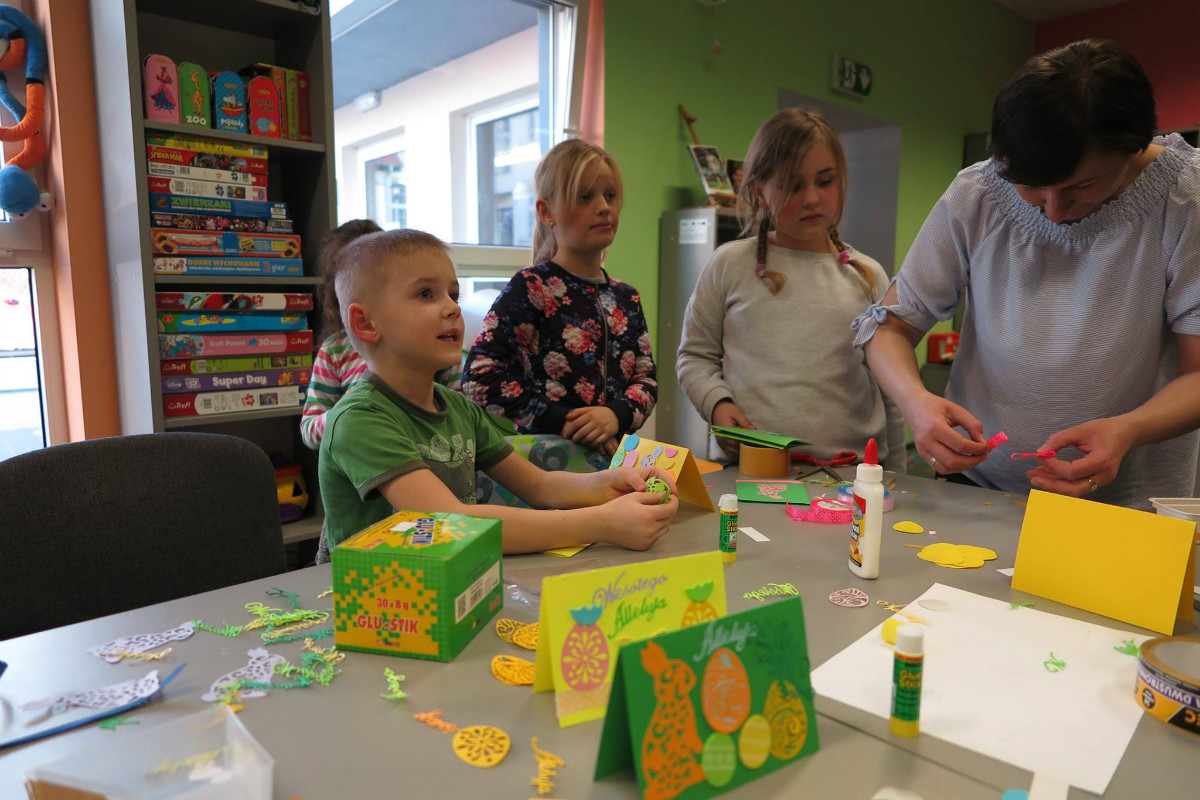 Wnętrze biblioteki. Przy stoliku czworo dzieci ozdabia kartki świąteczne pomaga im bibliotekarka Ania Wiśniewska. 