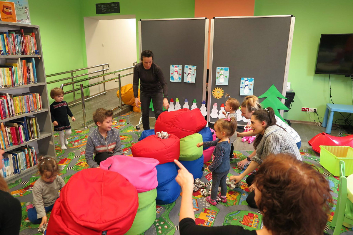 Na kolorowej wykładzinie siedzą dzieci z opiekunkami razem układają papierowe kulki i rzucają do celu. 
