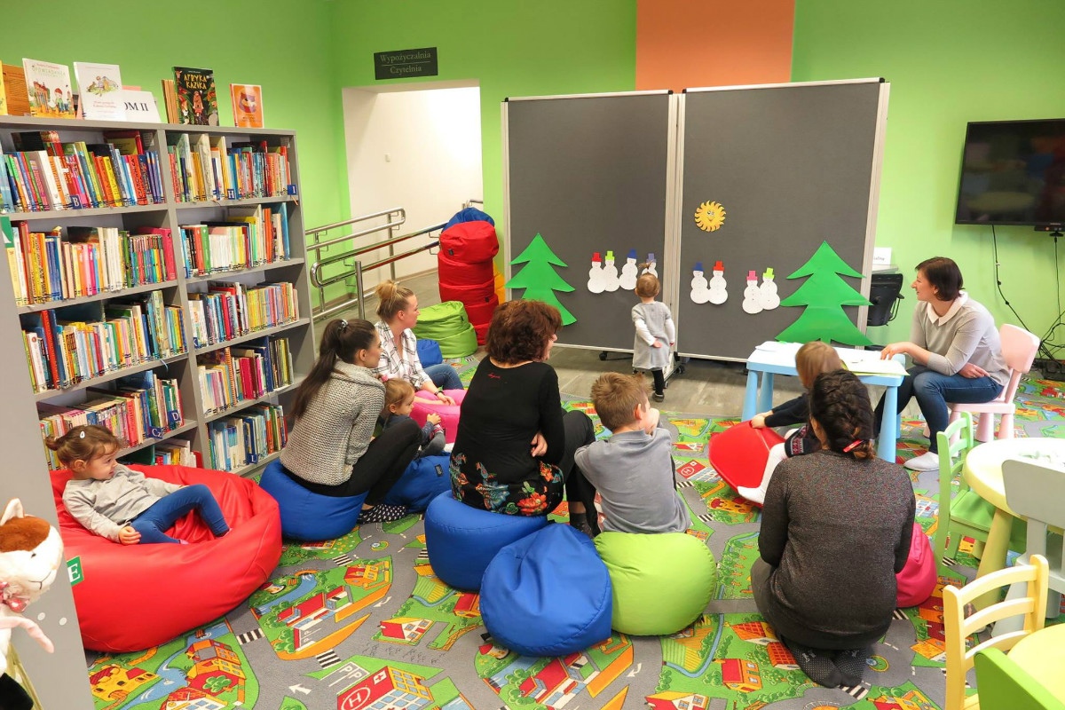Wnętrze biblioteki. Na pufach siedzą dzieci z opiekunkami, obok regały z książkami. Przy tablicy dziewczynka przygląda się bałwankom, zajęcia prowadzi bibliotekarka Ania Wiśniewska. 