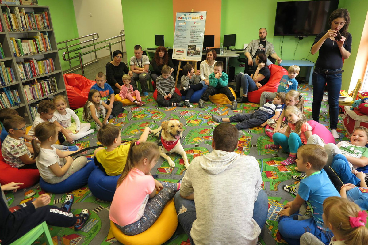 Wnętrze biblioteki. Na kolorowych pufach siedzą dzieci biorą udział w spotkaniu  z Panem Gracjanem Cichockim - dogoterpeutą w tle sztaluga z plakatem ''Zabajkowani 2''. 