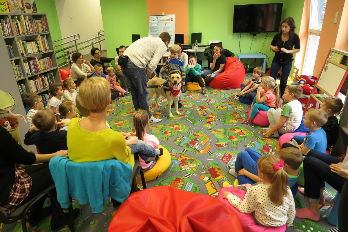 Wnętrze biblioteki. Na kolorowej wykładzinie siedzą dzieci, zajęcia prowadzi Gracjan Cichocki.