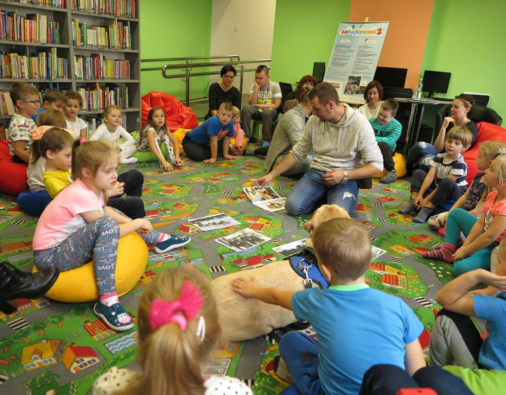 Wnętrze biblioteki. Na kolorowej wykładzinie siedzą dzieci, zajęcia prowadzi Gracjan Cichocki.