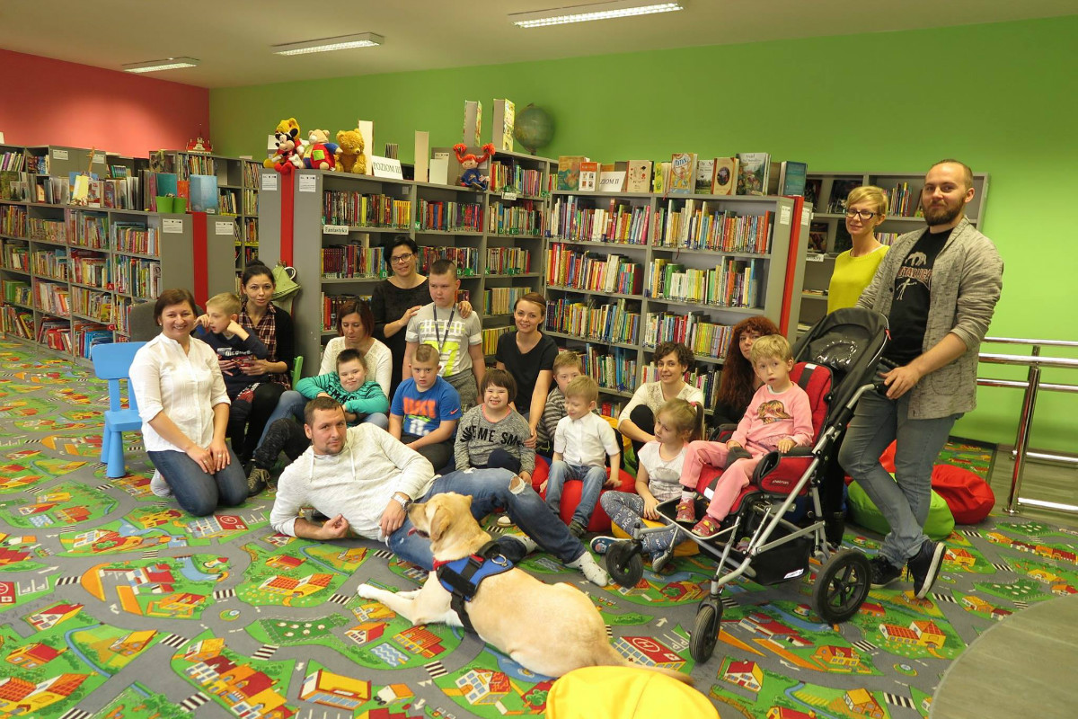 Wnętrze biblioteki. Na kolorowej wykładzinie siedzą dzieci i ich opiekunowie w tle regał z książkami.