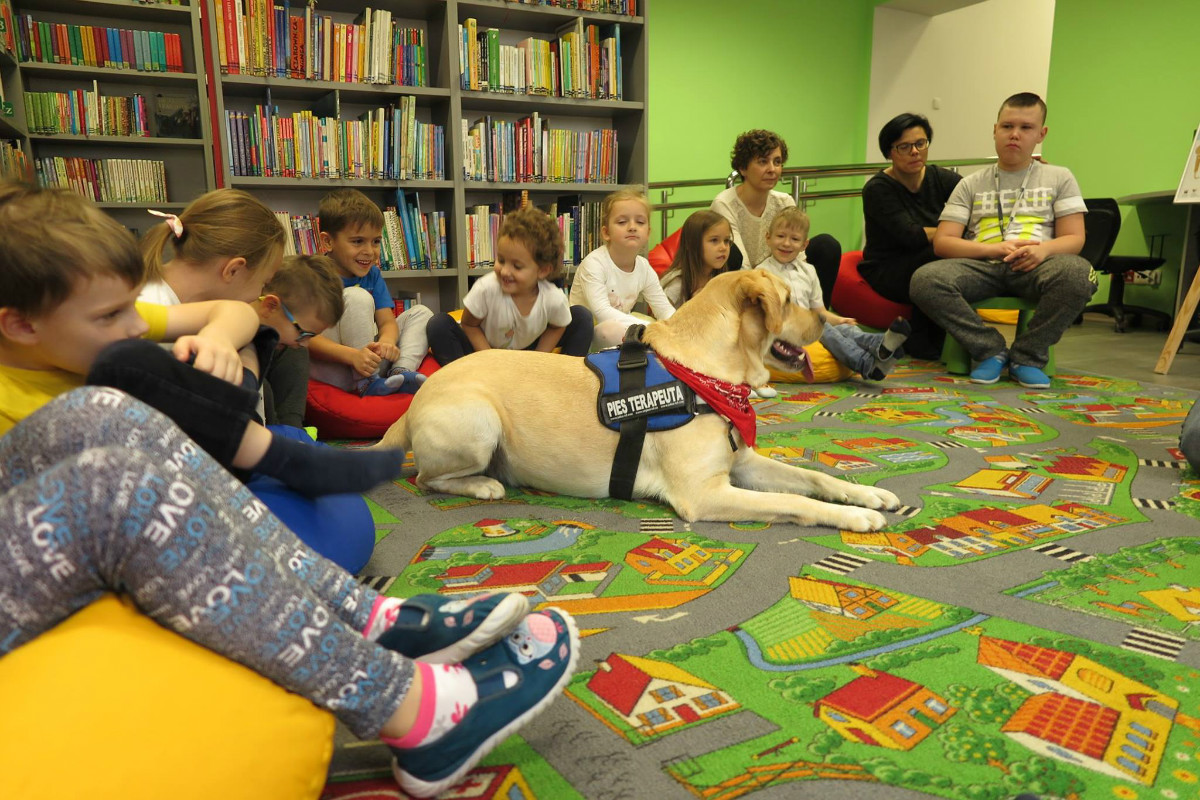 Wnętrze biblioteki. Na kolorowej wykładzinie siedzą dzieci obok nich pies labrador, zajęcia prowadzi Gracjan Cichocki.
