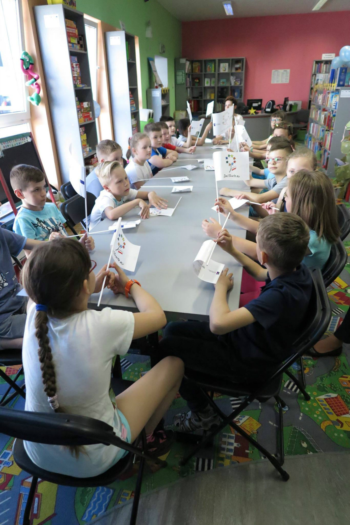 Przy stolikach siedzą dzieci, w dłoniach trzymają flagi z symbolem Dni Otwarte Unii Europejskiej. 