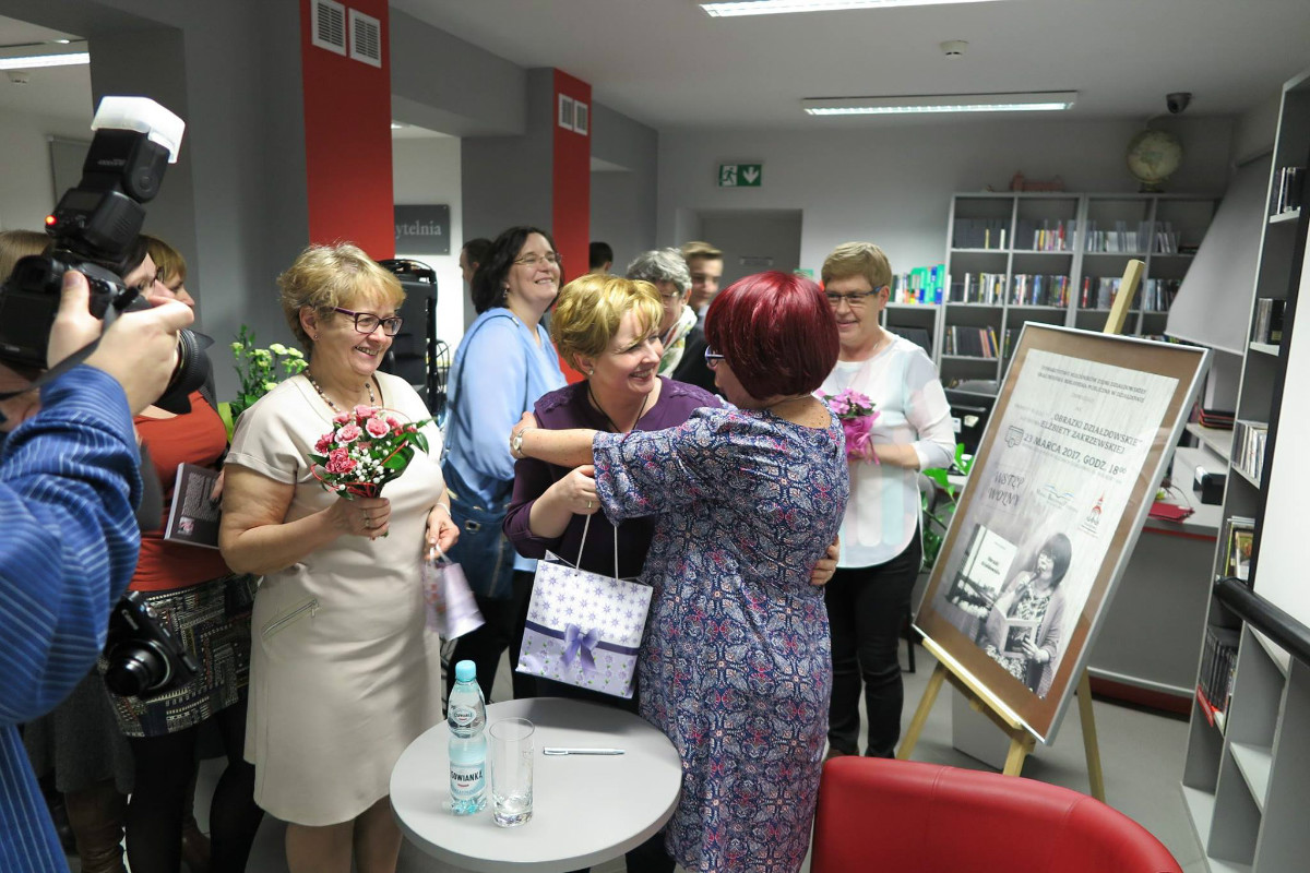 Wnętrze biblioteki,mediateka. Goście zgromadzeni na spotkanie wręczają kwiaty autorce Elżbiecie Zakrzewskiej. 