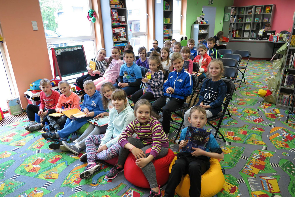 Na krzesełkach siedzą dzieci biorą udział w zajęciach w bibliotece. 