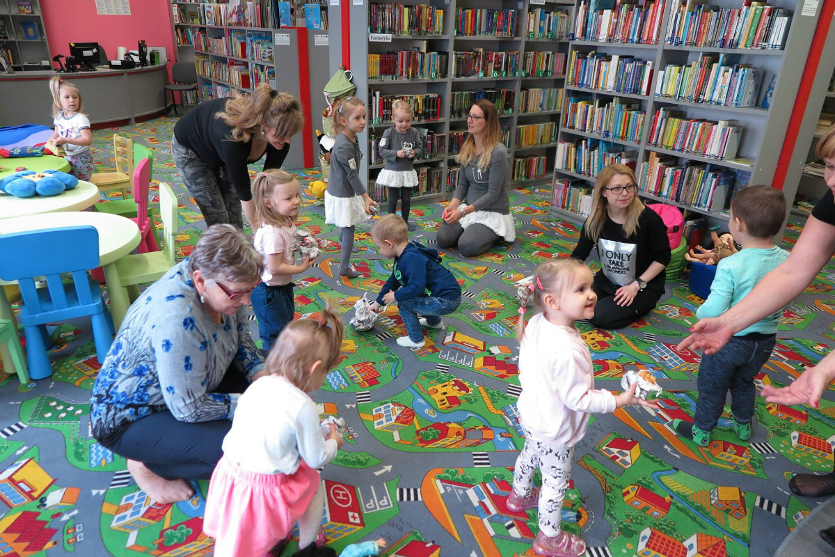 Wnętrze biblioteki. Na kolorowej wykładzinie bawią się dzieci z opiekunami podczas zajęć w klubie malucha.