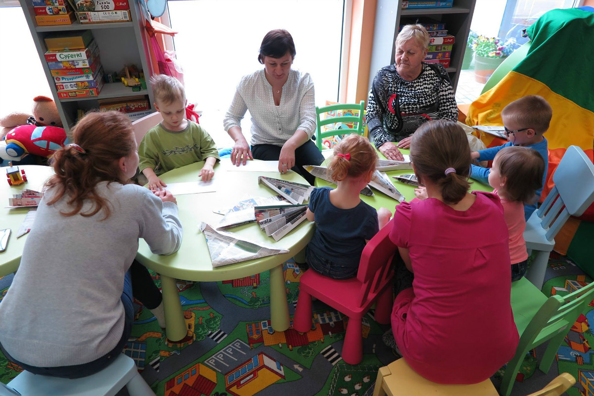 Przy stolikach siedzą dzieci z opiekunkami i bibliotekarką Anią Wiśniewską razem wykonują papierowe czapki z papieru. 