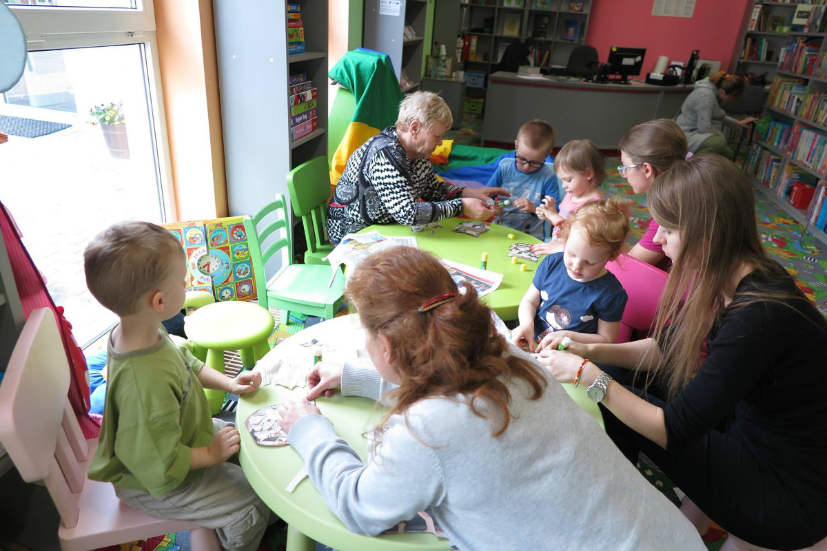 Przy stolikach siedzą dzieci z opiekunkami i bibliotekarką Olą Cybulską razem wykonują papierowe czapki z papieru. 