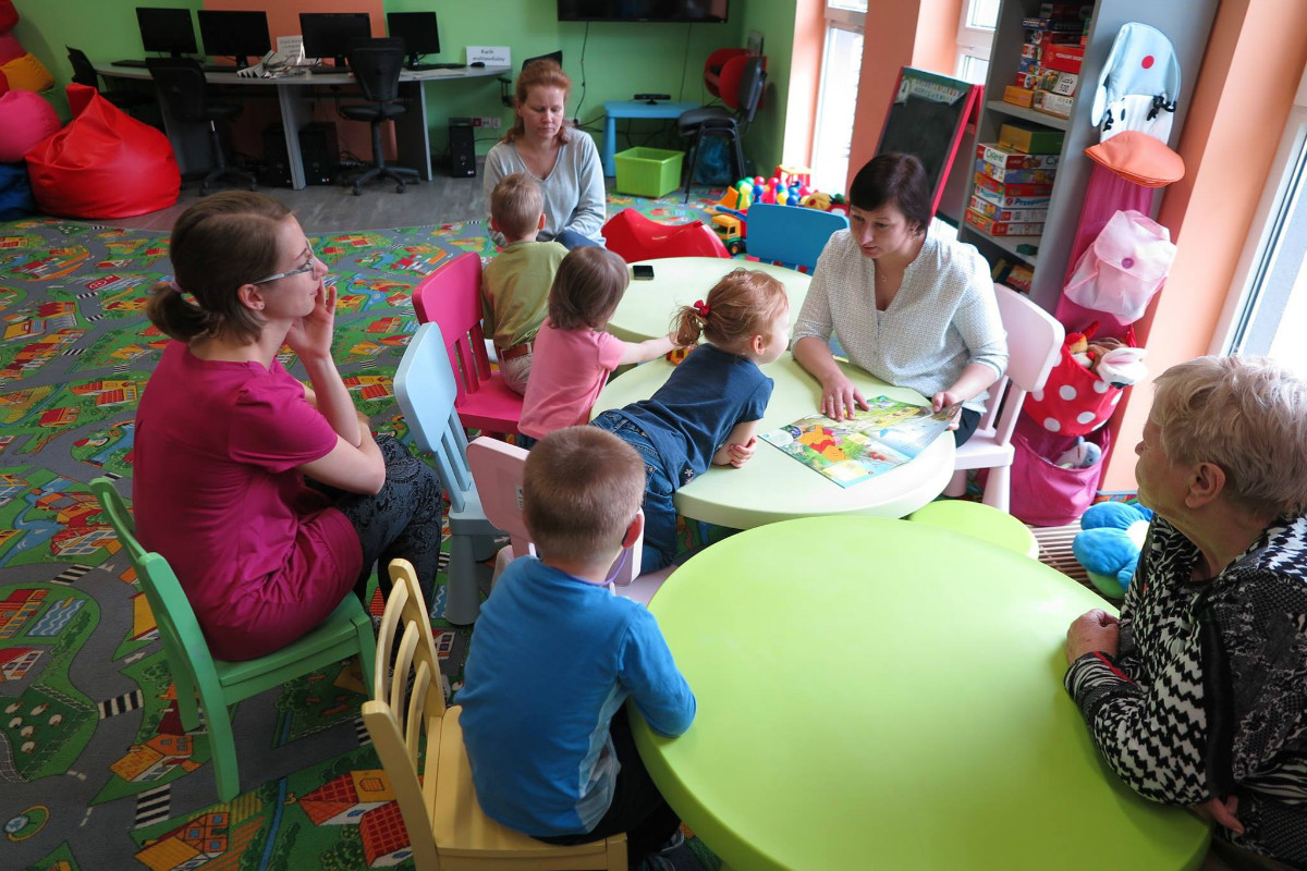 Przy stolikach siedzą dzieci z opiekunkami, bibliotekarka Ania Wiśniewska pokazuje dzieciom gazetkę z Kubusiem Puchatkiem. 