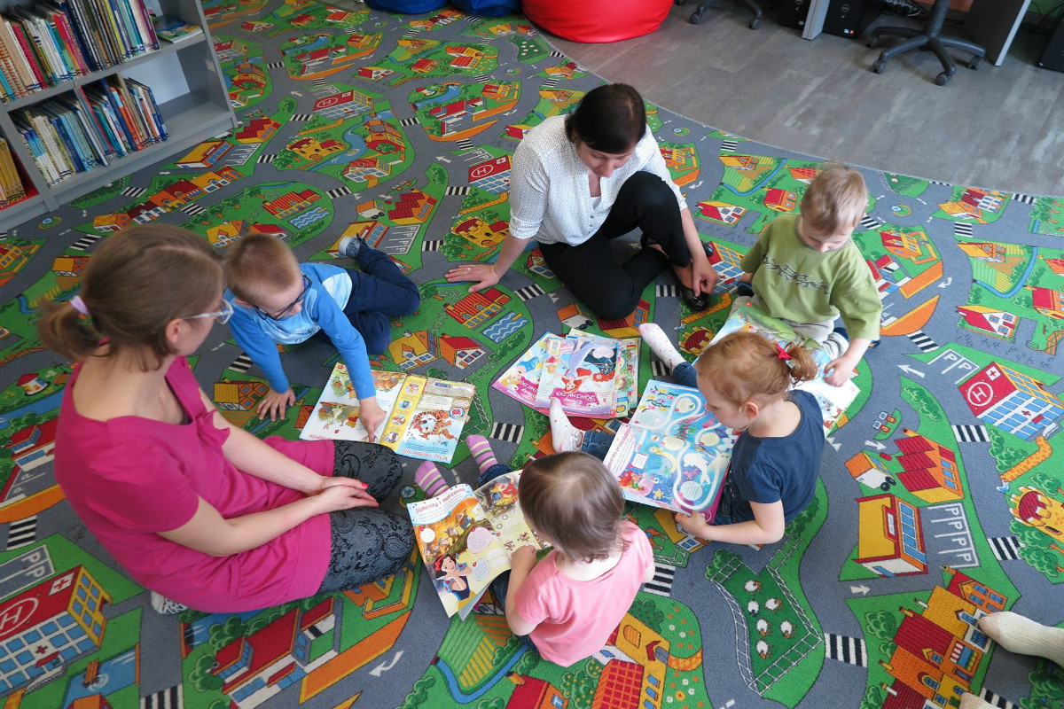 Wnętrze biblioteki. Na kolorowej wykładzinie siedzą dzieci z bibliotekarką Anią Wiśniewską i przeglądają gazetki dla dzieci. 