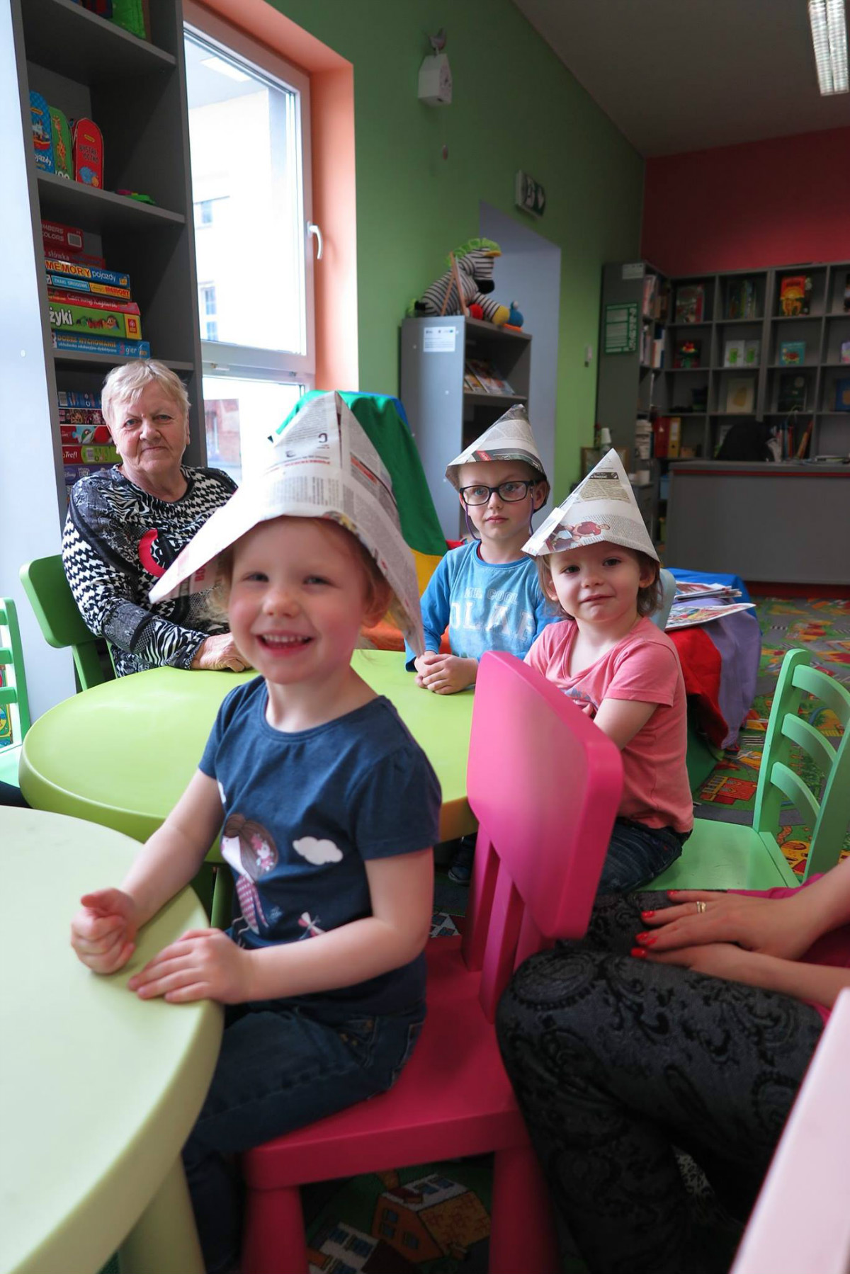 Przy stoliku siedzi opiekunka i troje dzieci z papierowymi czapkami na głowie. 