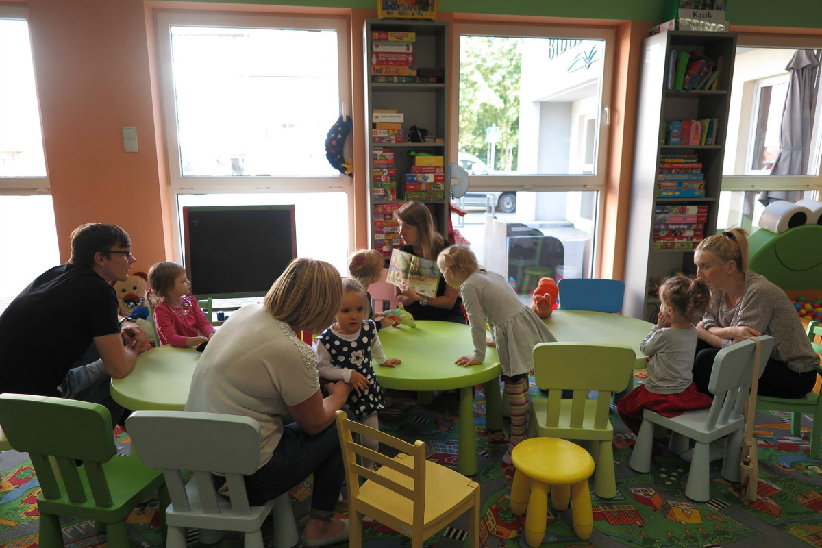 Wnętrze biblioteki. Przy  stolikach siedzą dzieci z opiekunami, bibliotekarka Aleksandra Cybulska prowadzi zajęcia. 