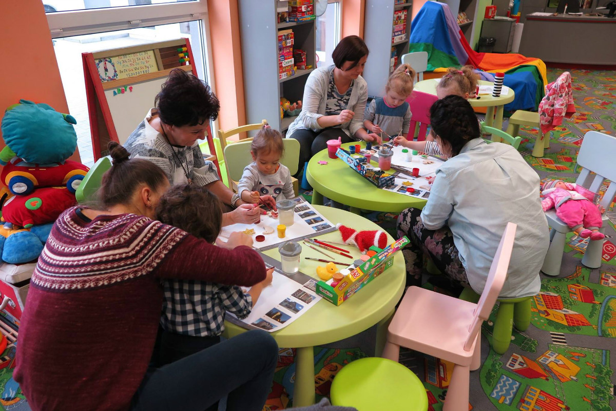Wnętrze biblioteki. Przy stolikach siedzą dzieci z opiekunami wspólnie malują farbami ozdoby choinkowe.