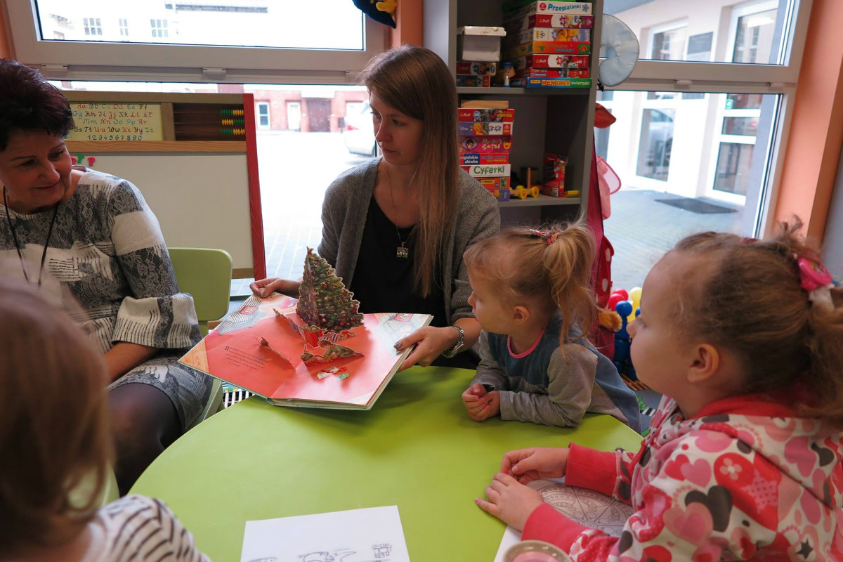 Wnętrze biblioteki. Przy stoliku siedzą dzieci z opiekunami, bibliotekarka Aleksandra Cybulska wspólnie z dziećmi ogląda i czyta  książkę "Prezent dla Świętego Mikołaja". 