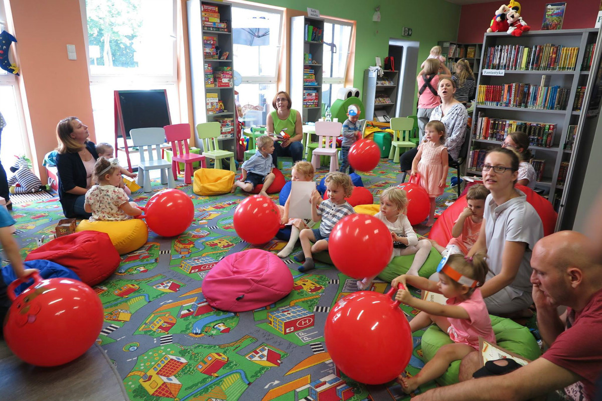 Wnętrze biblioteki. Na czerwonej wykładzinie siedzą dzieci z czerwonymi piłkami i ich opiekunowie. 