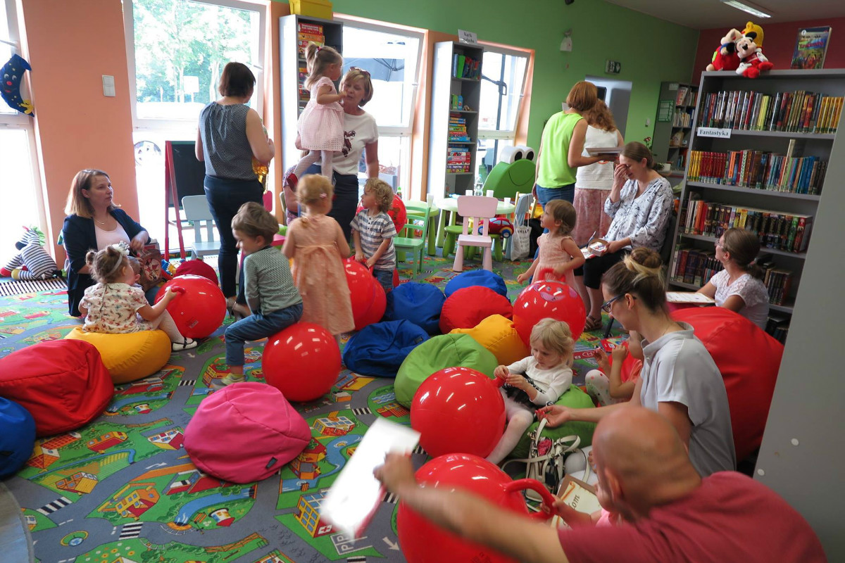Wnętrze biblioteki. Na czerwonej wykładzinie siedzą dzieci z czerwonymi piłkami i ich opiekunowie. 