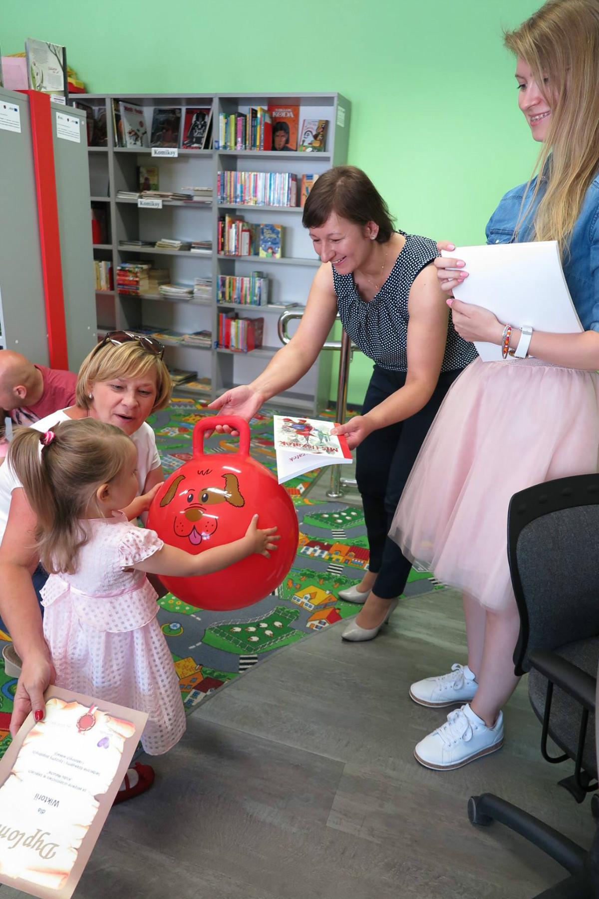 Bibliotekarka Ania Wiśniewska wręcza nagrodę dziewczynce czerwoną piłkę.