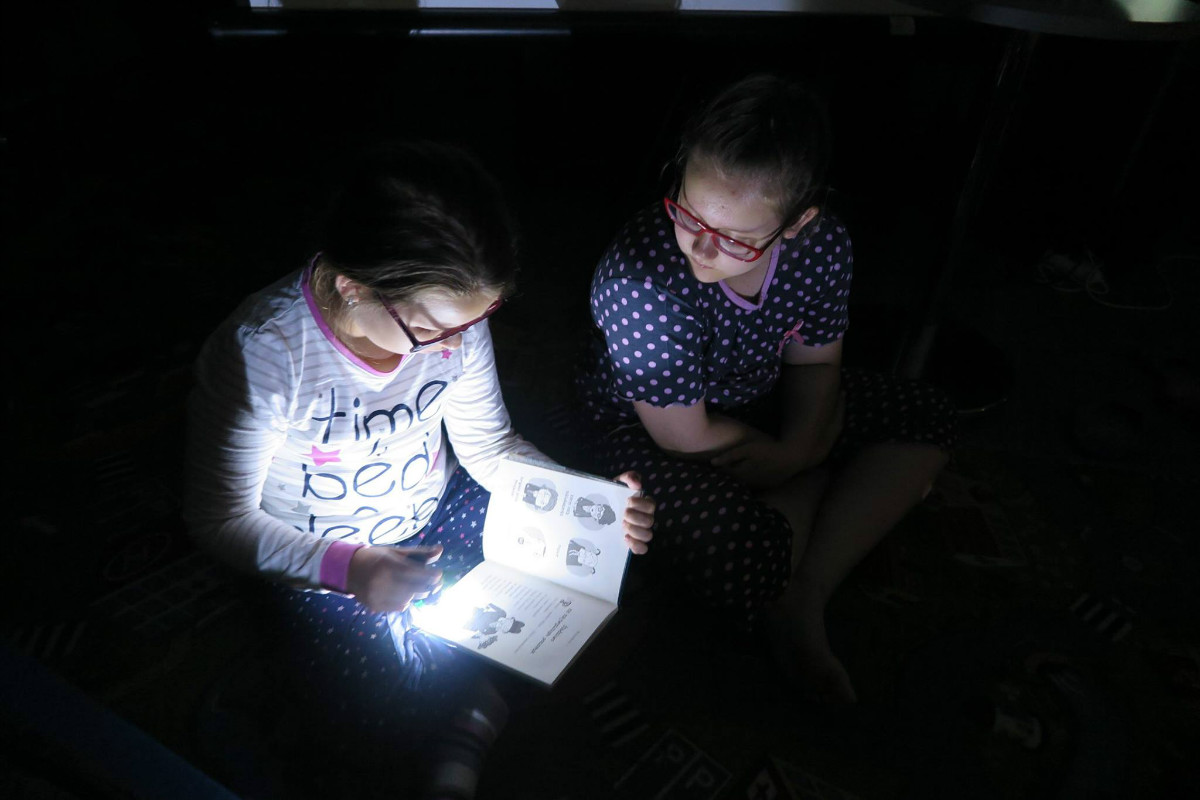 Wnętrze biblioteki. Podczas nocy w bibliotece dwie dziewczynki czytają książkę z pomocą latarki.