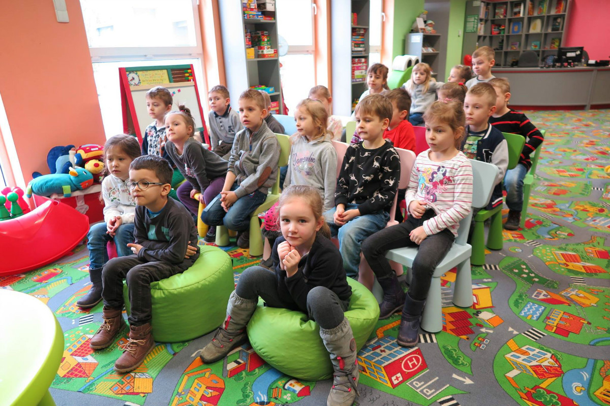 Wnętrze biblioteki, na krzesełkach siedzą dzieci z grupy III z Przedszkola nr 5 biorą udział w lekcji bibliotecznej. 