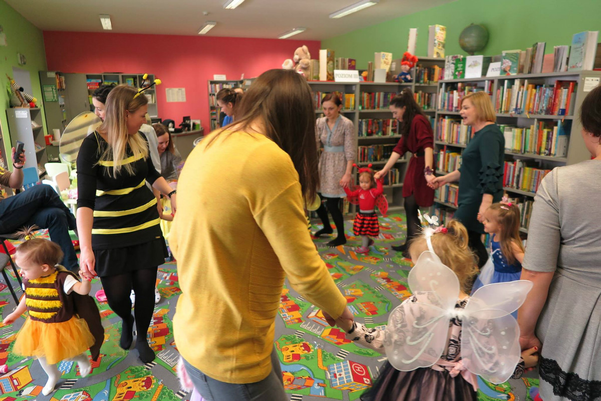 Wnętrze biblioteki. Dzieci przebrane w kolorowe stroje tańczą w kółku z opiekunami podczas balu karnawałowego.