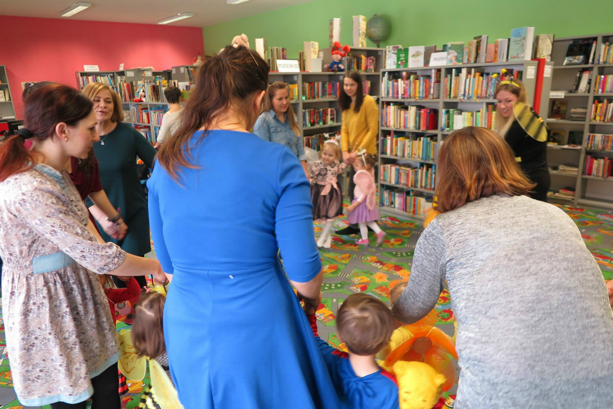 Wnętrze biblioteki. Dzieci przebrane w kolorowe stroje tańczą w kółku z opiekunami podczas balu karnawałowego.