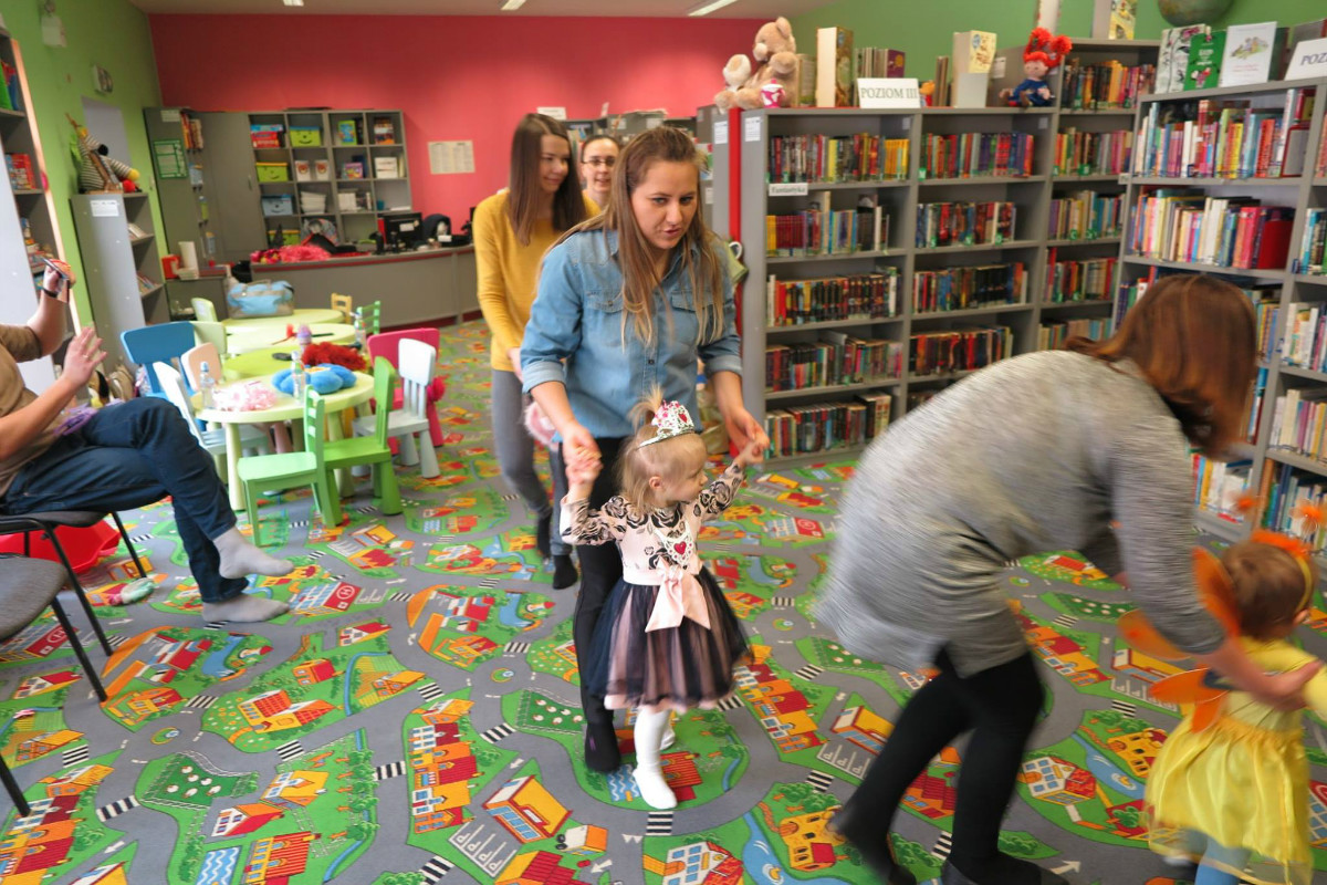 Wnętrze biblioteki. Dzieci przebrane w kolorowe stroje tańczą z opiekunami podczas balu karnawałowego.