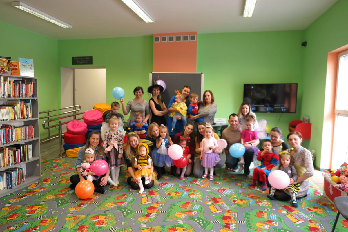 Wnętrze biblioteki. Grupa dzieci przebrana w kolorowe stroje trzyma balony z okazji balu karnawałowego.