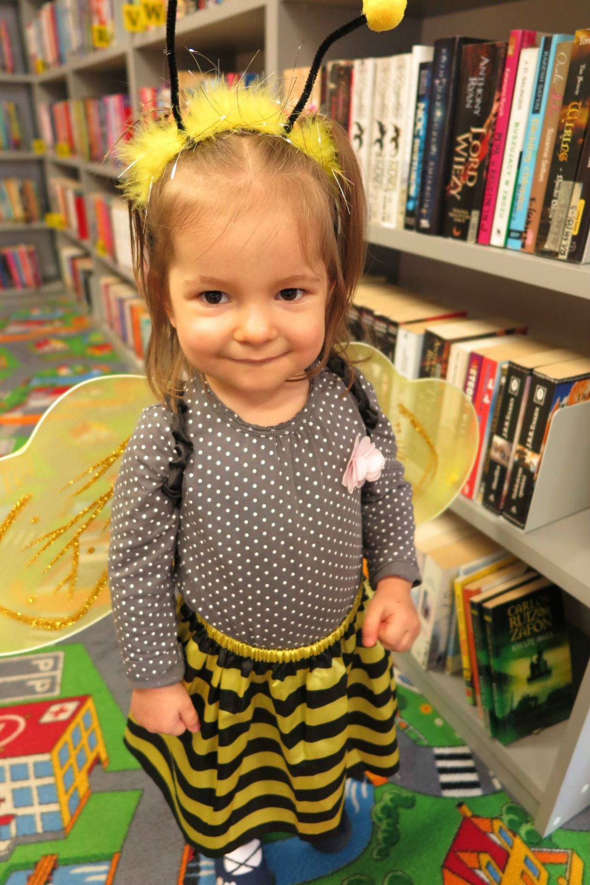 Przy regale z książkami stoi dziewczynka w stroju pszczółki w trakcie balu.