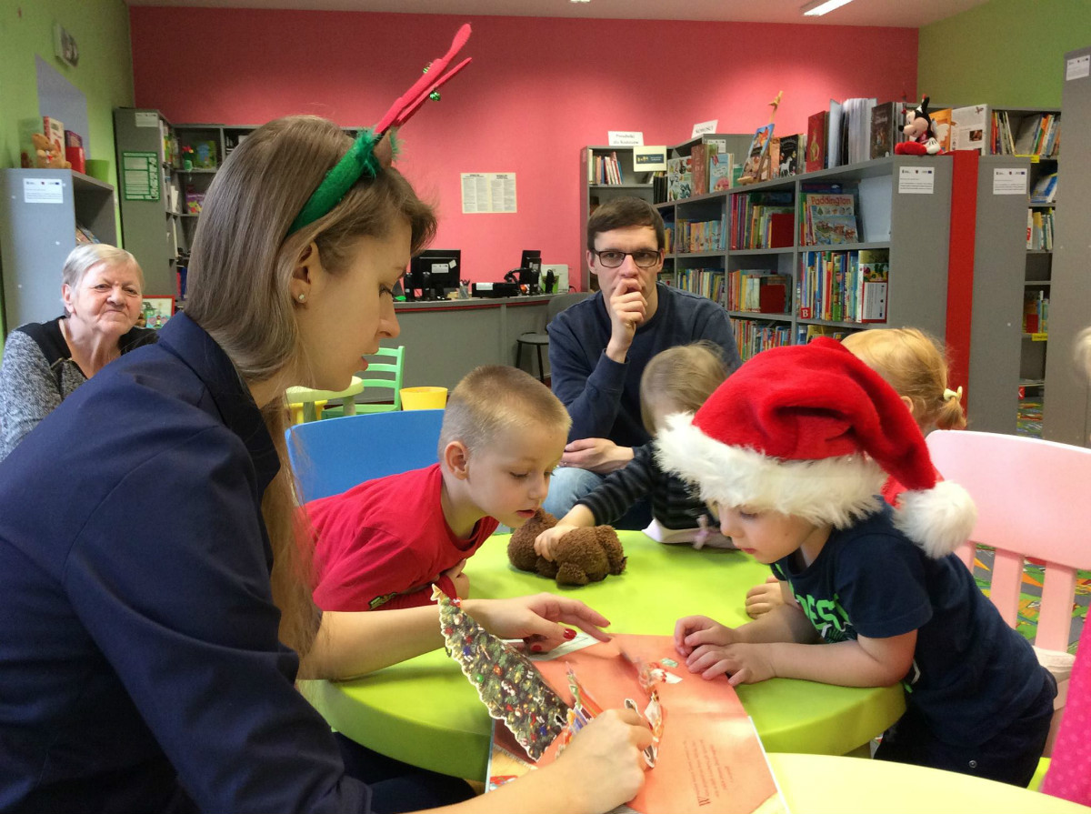 Wnętrze biblioteki, bibliotekarka Aleksandra Cybulska razem z dziećmi ogląda trójwymiarową książeczkę "Prezent dla Świętego Mikołaja". 