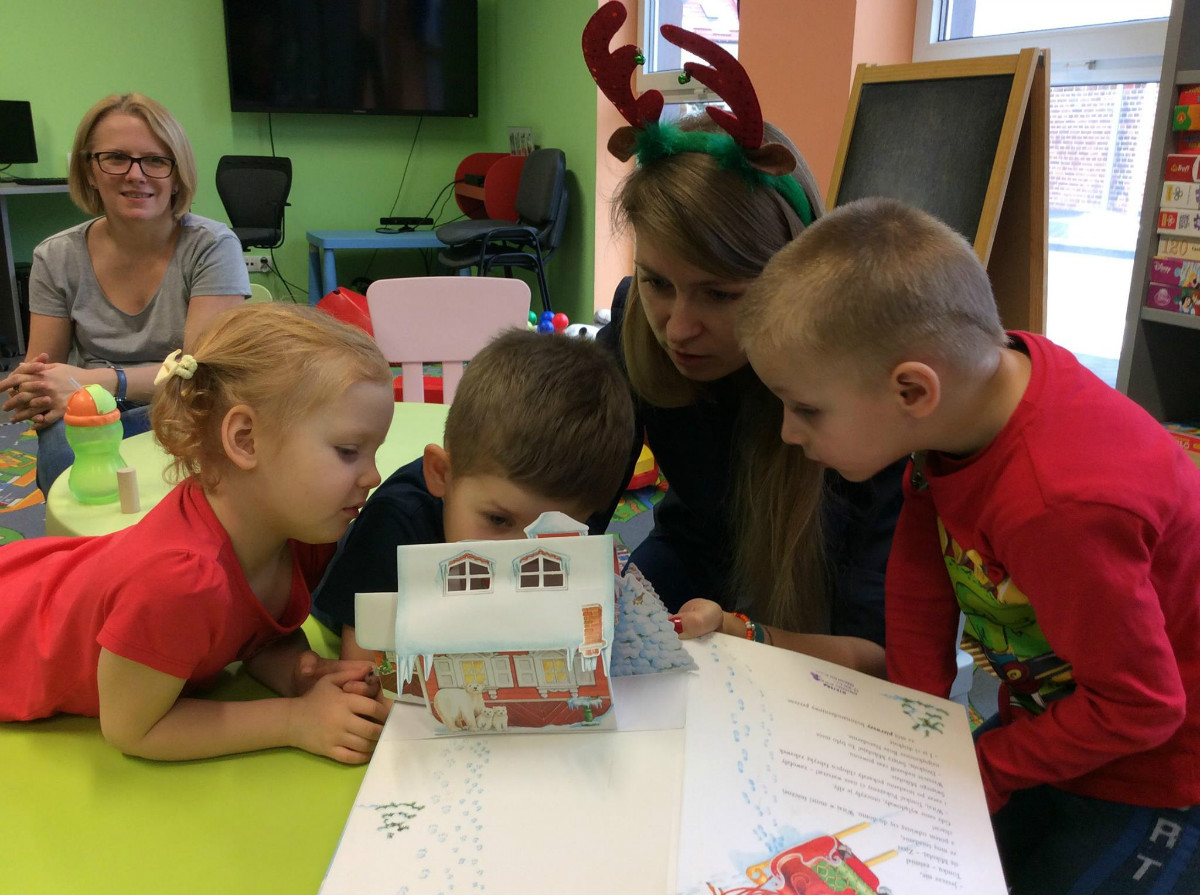 Wnętrze biblioteki, bibliotekarka Aleksandra Cybulska razem z dziećmi ogląda trójwymiarową książeczkę "Prezent dla Świętego Mikołaja". 