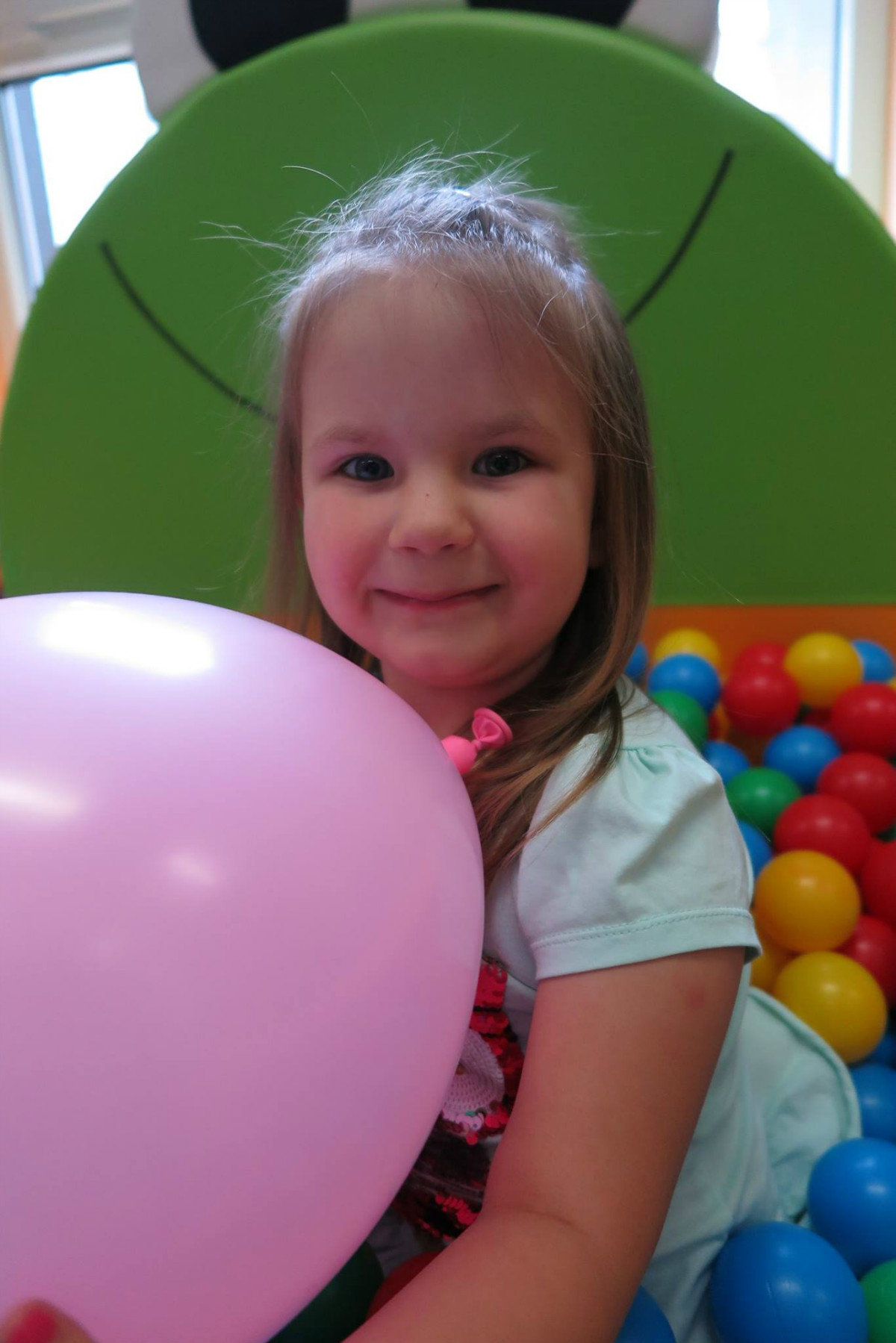 W basenie z kolorowymi piłeczkami siedzi dziewczynka z balonem.