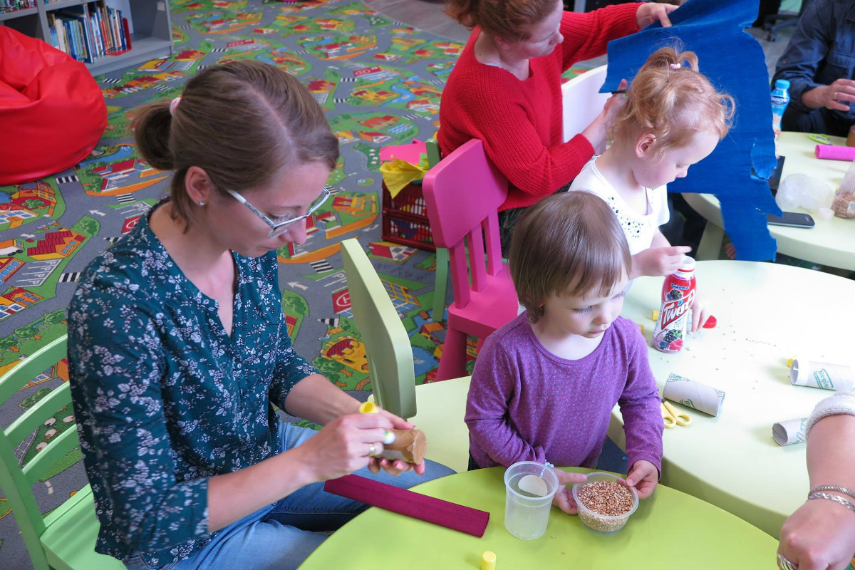 Przy stolikach siedzą dzieci z opiekunami razem wykonują prace plastyczne.