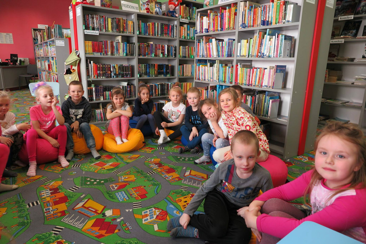 Wnętrze biblioteki. Przy regale z książkami siedzą dzieci z Przedszkola nr 3 biorą udział w lekcji bibliotecznej. 