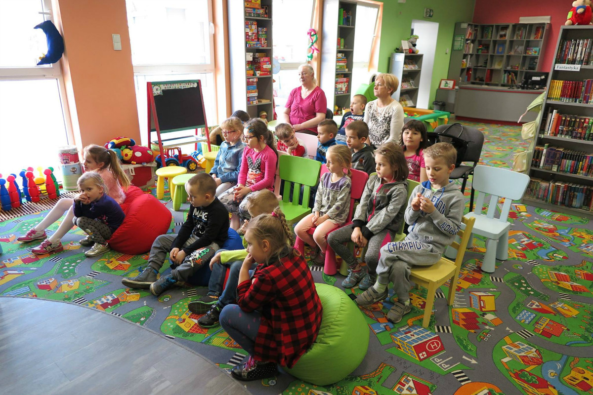 Wnętrze biblioteki. Na krzesełkach siedzą dzieci, bibliotekarka Ania Wiśniewska prowadzi zajęcia. 