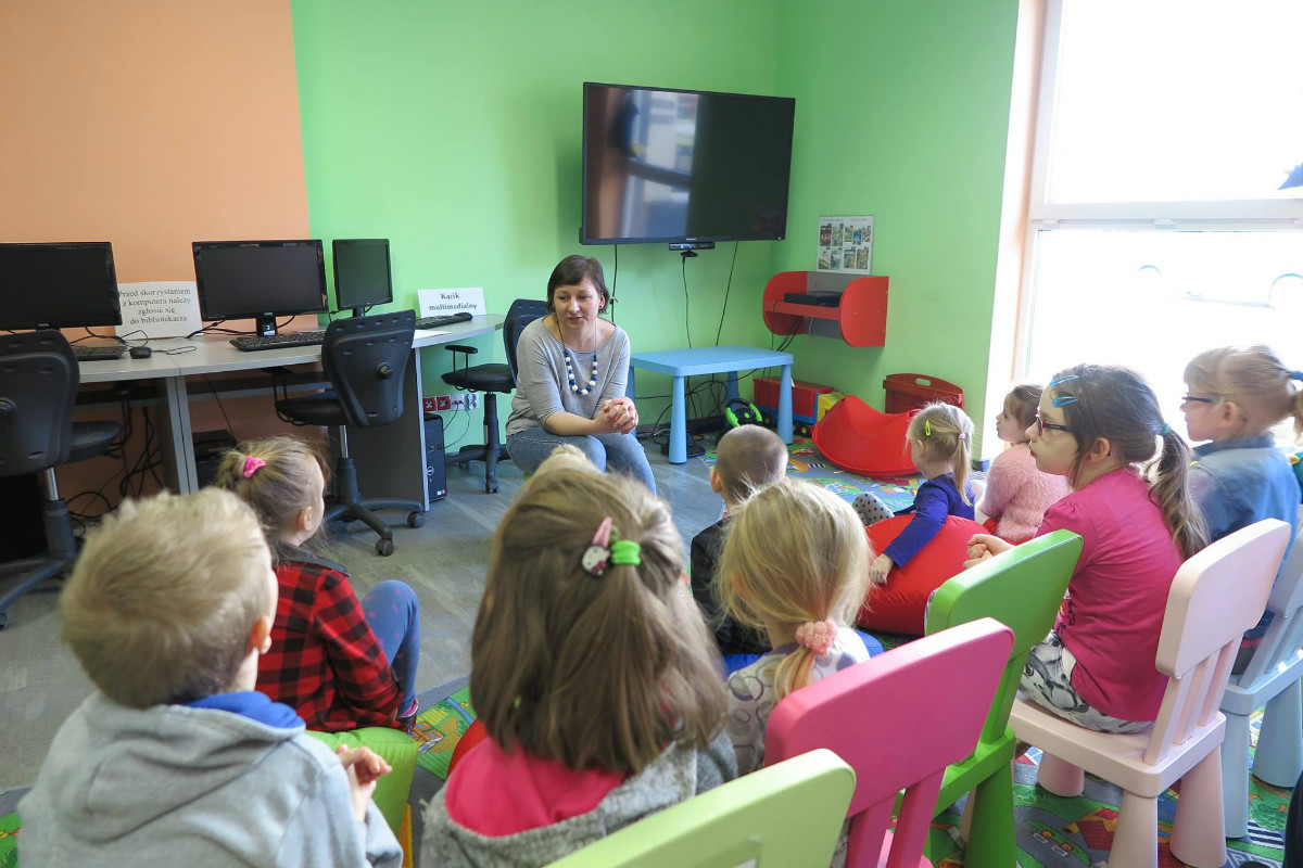 Wnętrze biblioteki. Na krzesełkach siedzą dzieci, bibliotekarka Ania Wiśniewska prowadzi zajęcia. 
