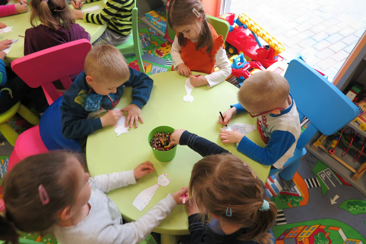 Wnętrze biblioteki. Przy stoliku siedzą dzieci wspólnie kolorują kredkami. 