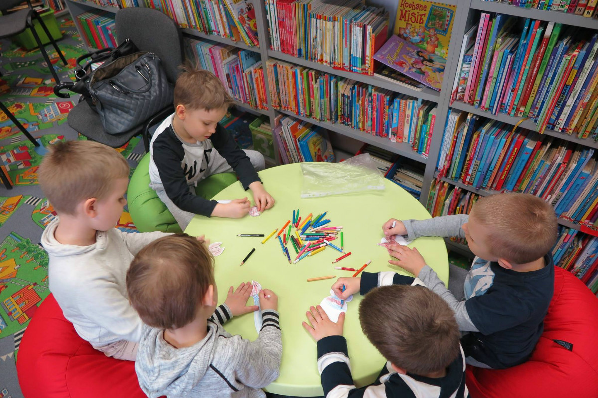 Wnętrze biblioteki. Przy stoliku siedzą chłopcy wspólnie kolorują kredkami. 