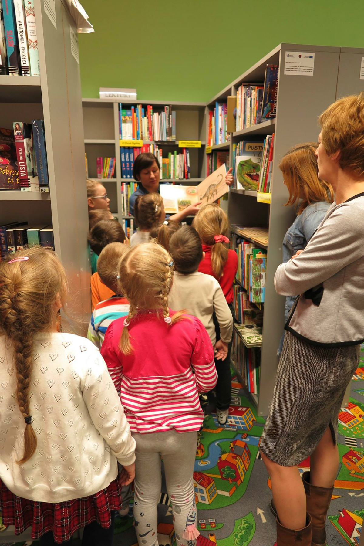 Między regałami stoi grupa dzieci, bibliotekarka pokazuje im książkę o dinozaurach. 