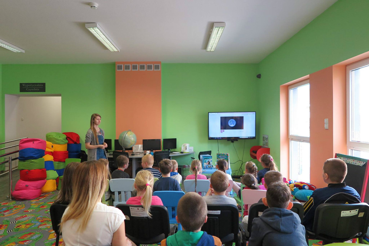 Wnętrze biblioteki. Dzieci wraz z bibliotekarką Aleksandrą Cybulską oglądają prezentację na temat obiegu wody w przyrodzie oraz jej oszczędzaniu.