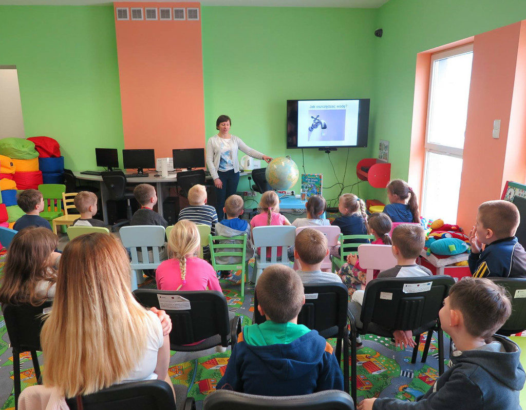 Wnętrze biblioteki. Dzieci wraz z bibliotekarką Anią Wiśniewską oglądają prezentację na temat obiegu wody w przyrodzie oraz jej oszczędzaniu.