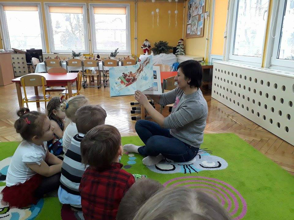 Wnętrze Przedszkola nr 4, na zielonym dywanie siedzą dzieci, bibliotekarka Ania Wiśniewska pokazuje i czyta  bożonarodzeniowe opowiadania "Prezent dla Świętego Mikołaja" i "W oczekiwaniu na Pierwszą Gwiazdkę".