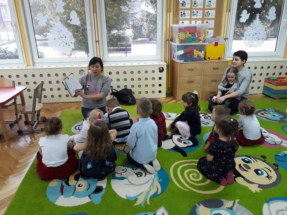 Wnętrze Przedszkola nr 4, na zielonym dywanie siedzą dzieci, bibliotekarka Ania Wiśniewska pokazuje i czyta  bożonarodzeniowe opowiadania "Prezent dla Świętego Mikołaja" i "W oczekiwaniu na Pierwszą Gwiazdkę".