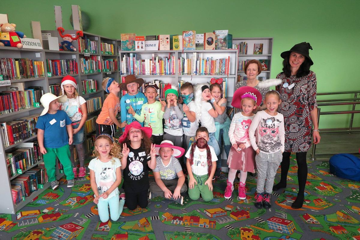 Wnętrze biblioteki, grupa dzieci biorąca udział w bajkowych warsztatach prowadzonych przez  pisarkę  Agatę Grzegorczyk-Wosiek, w tle regały z książkami.