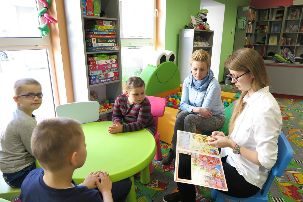 Wnętrze biblioteki. Bibliotekarka Aleksandra Cybulska czyta dzieciom opowiadanie "Trzy świnki".