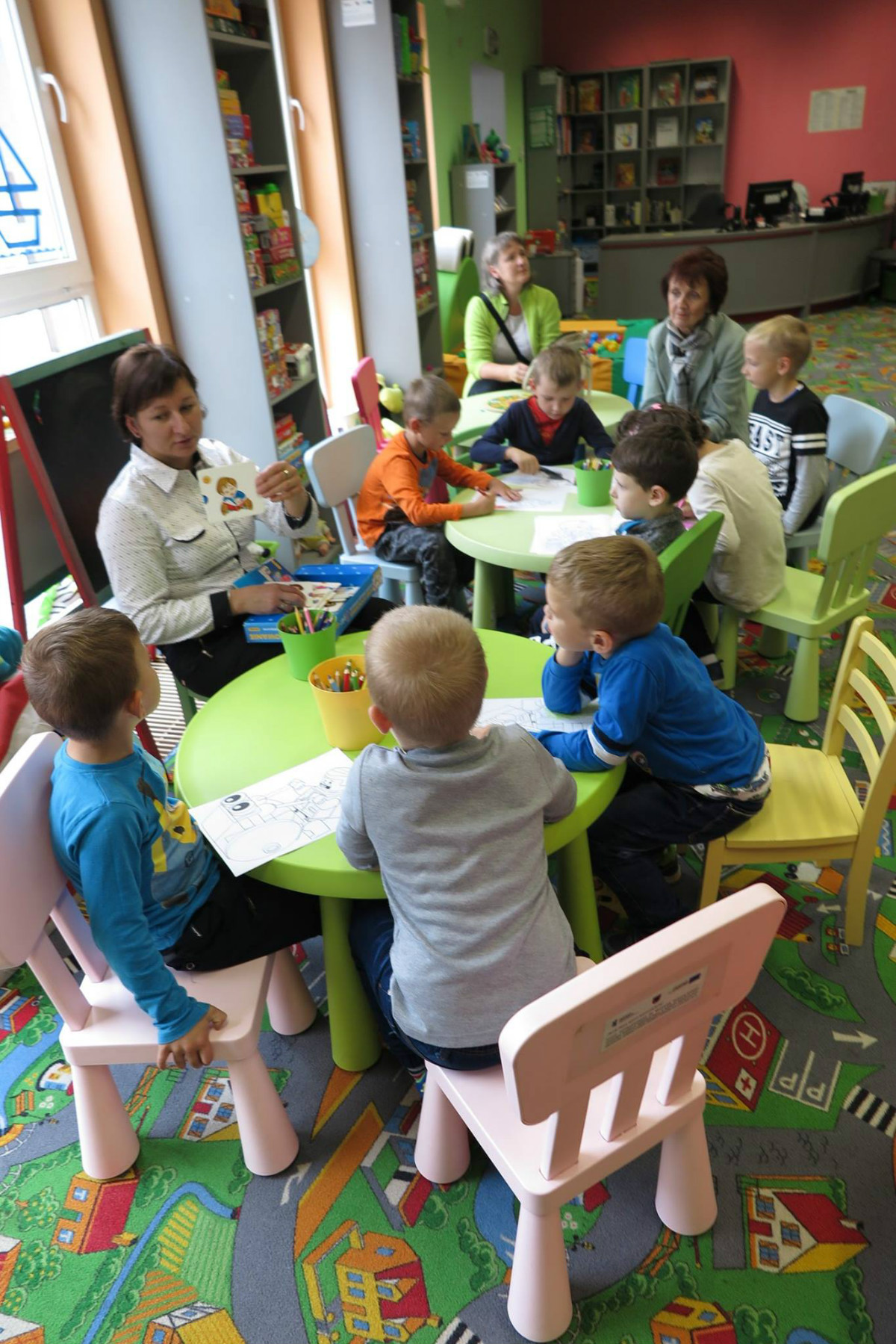 Wnętrze biblioteki, przy stolikach siedzą dzieci z opiekunami, bibliotekarka Ania Wiśniewska prowadzi zajęcia. 
